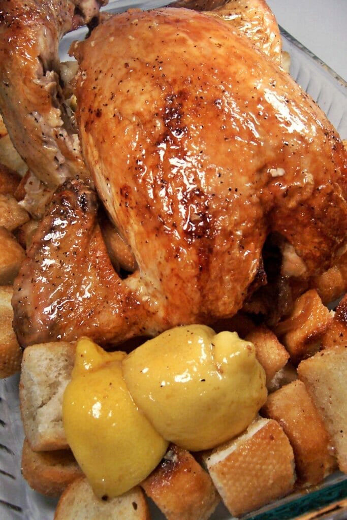 A browned, roasted whole chicken with a lemon glaze on top. Surrounded by large pieces of croutons made from a fresh baguette and halved lemons.