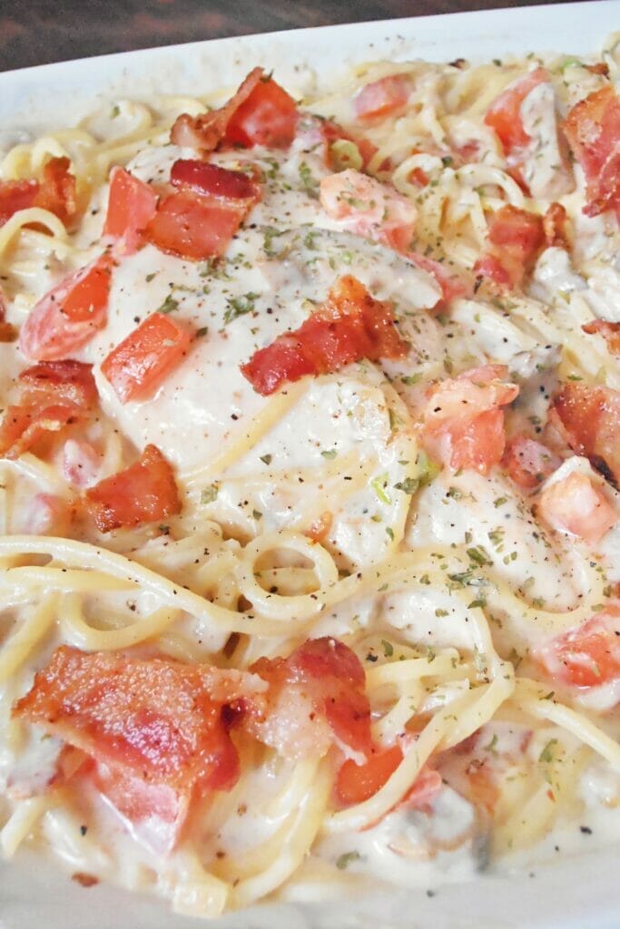 Close up of cooked angel hair pasta that has been tossed with a creamy, cheesy Caesar based sauce. Topped with chicken breasts, parsley, chopped tomatoes and bacon.