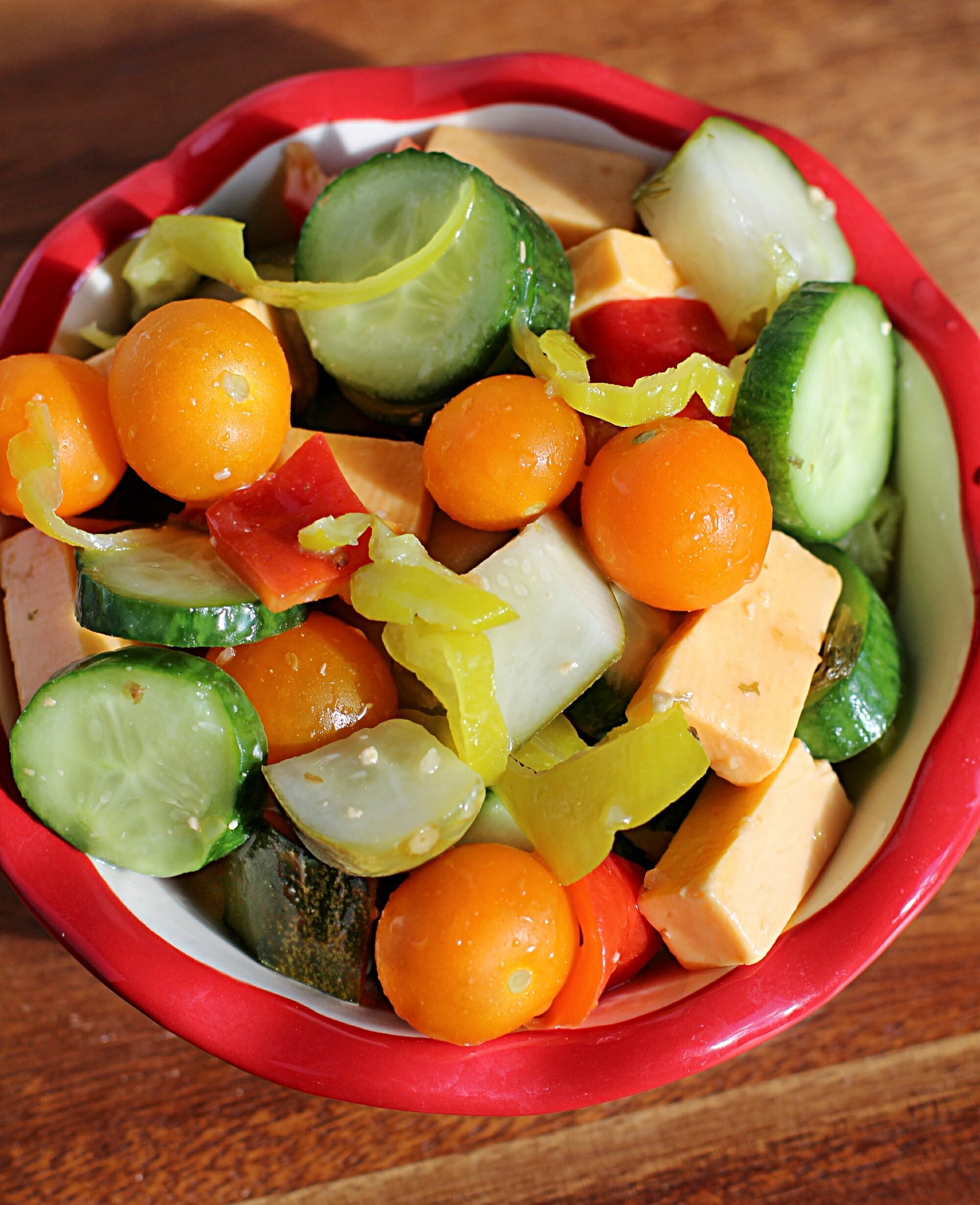 A red and white side dish is filled with chopped cucumbers, pickles, sliced pepperoncini, cubed cheddar cheese and ripe cherry tomatoes.